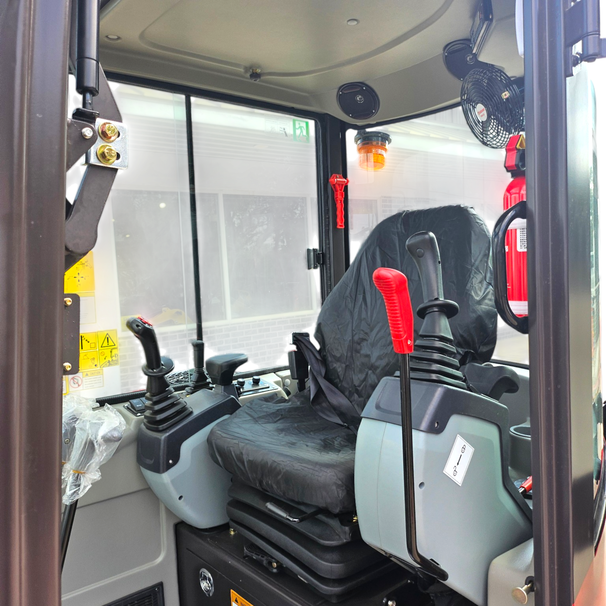 Interior of a truck cab showing the driver's seat and control levers, with an orange warning beacon on the console and a large fan on the wall; emergency exit sign visible through the window.