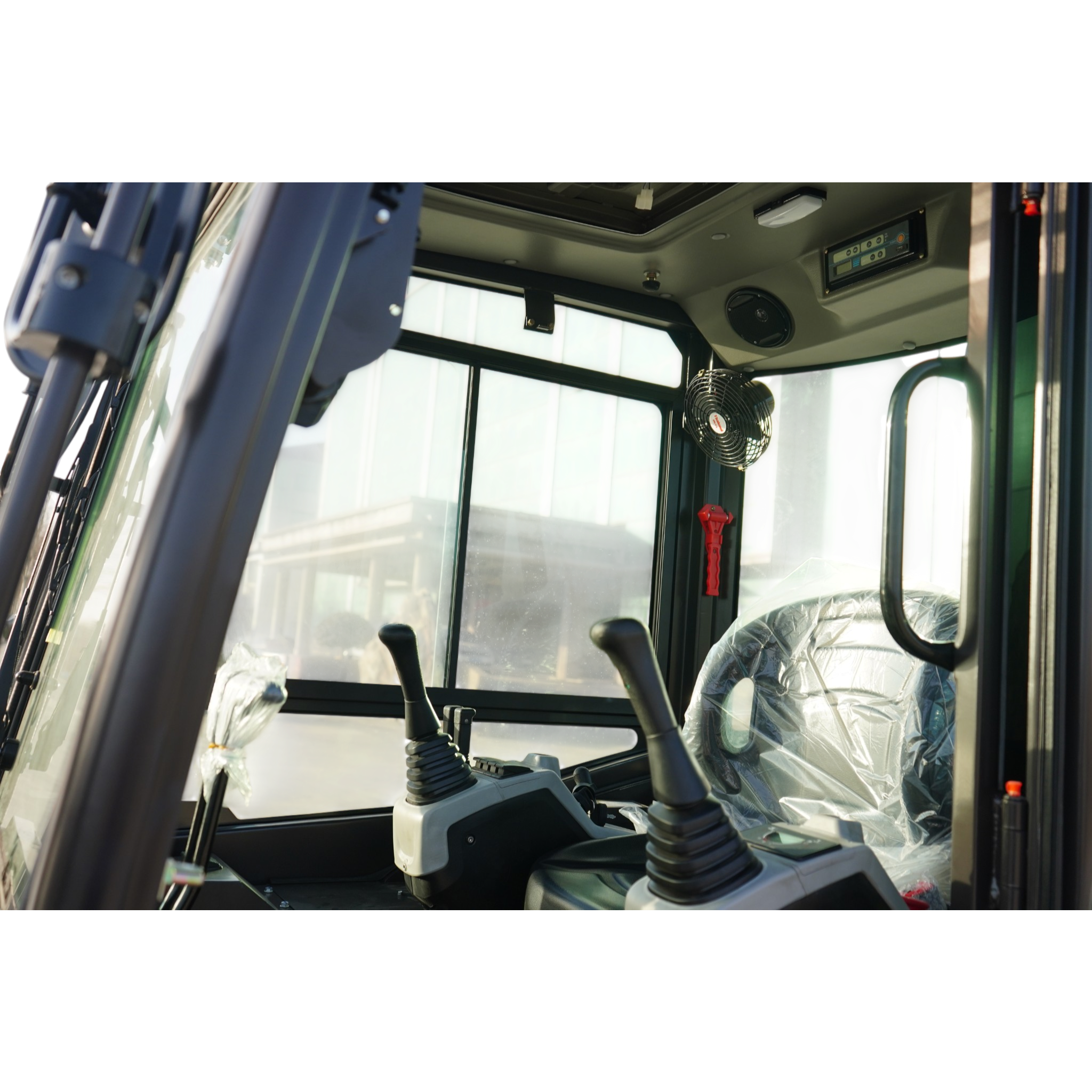 Interior view of a construction vehicle cab with multiple control levers, a digital display, and a round vent fan visible near the window edge.