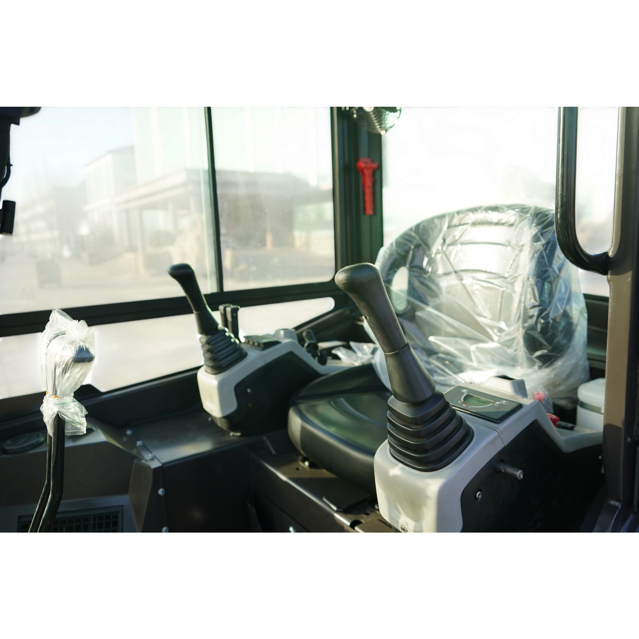 Cab interior of a heavy equipment vehicle with two joystick control levers and a plastic-wrapped seat, viewed through the windshield and windows