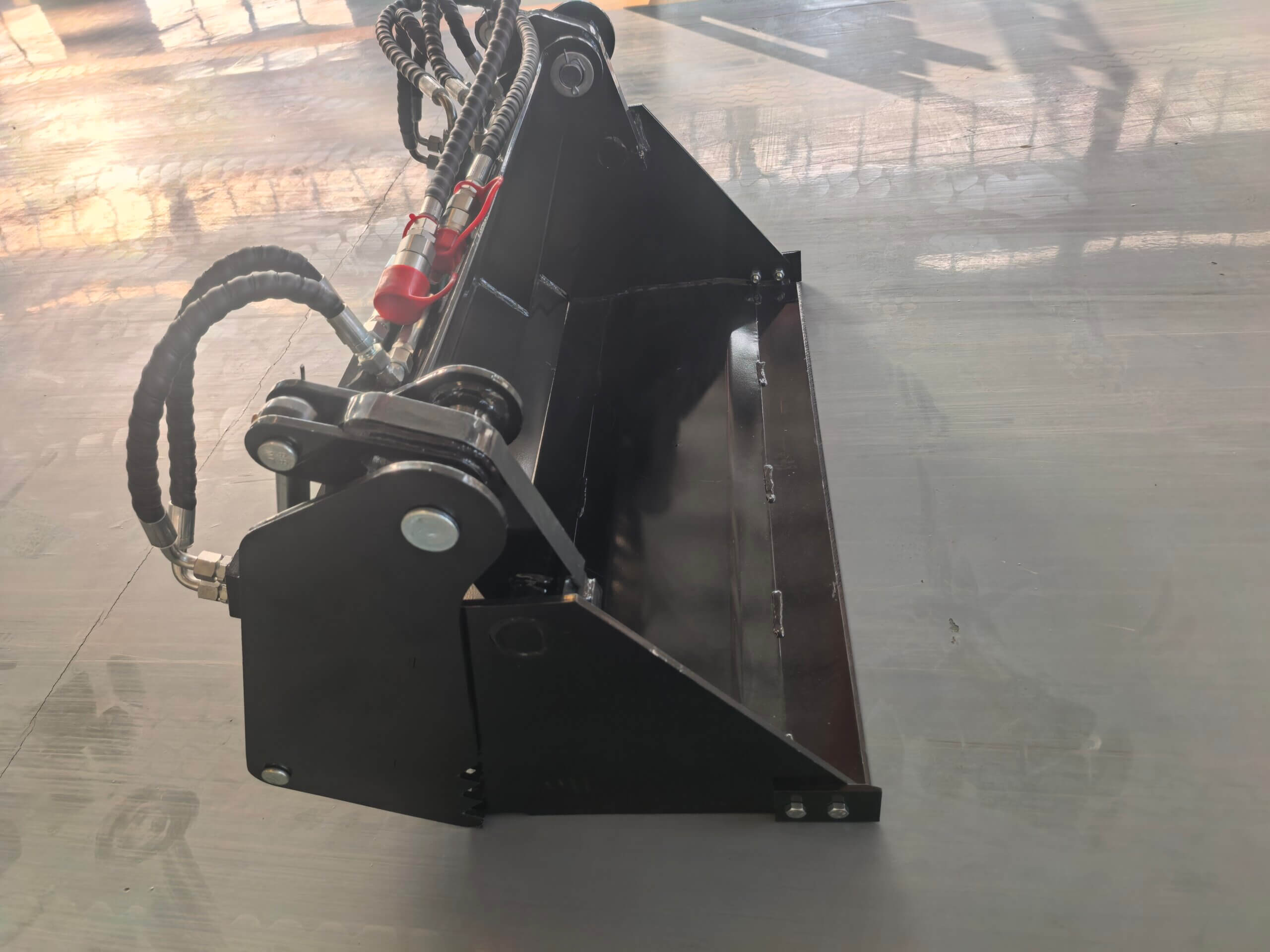 Industrial hydraulic bucket attachment with green hoses and red quick-connect fitting and pivoting joints, mounted on a black metal frame.