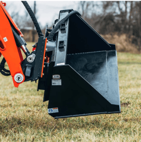 Black bucket attachment of a construction excavator on a grassy field, ready to scoop soil.