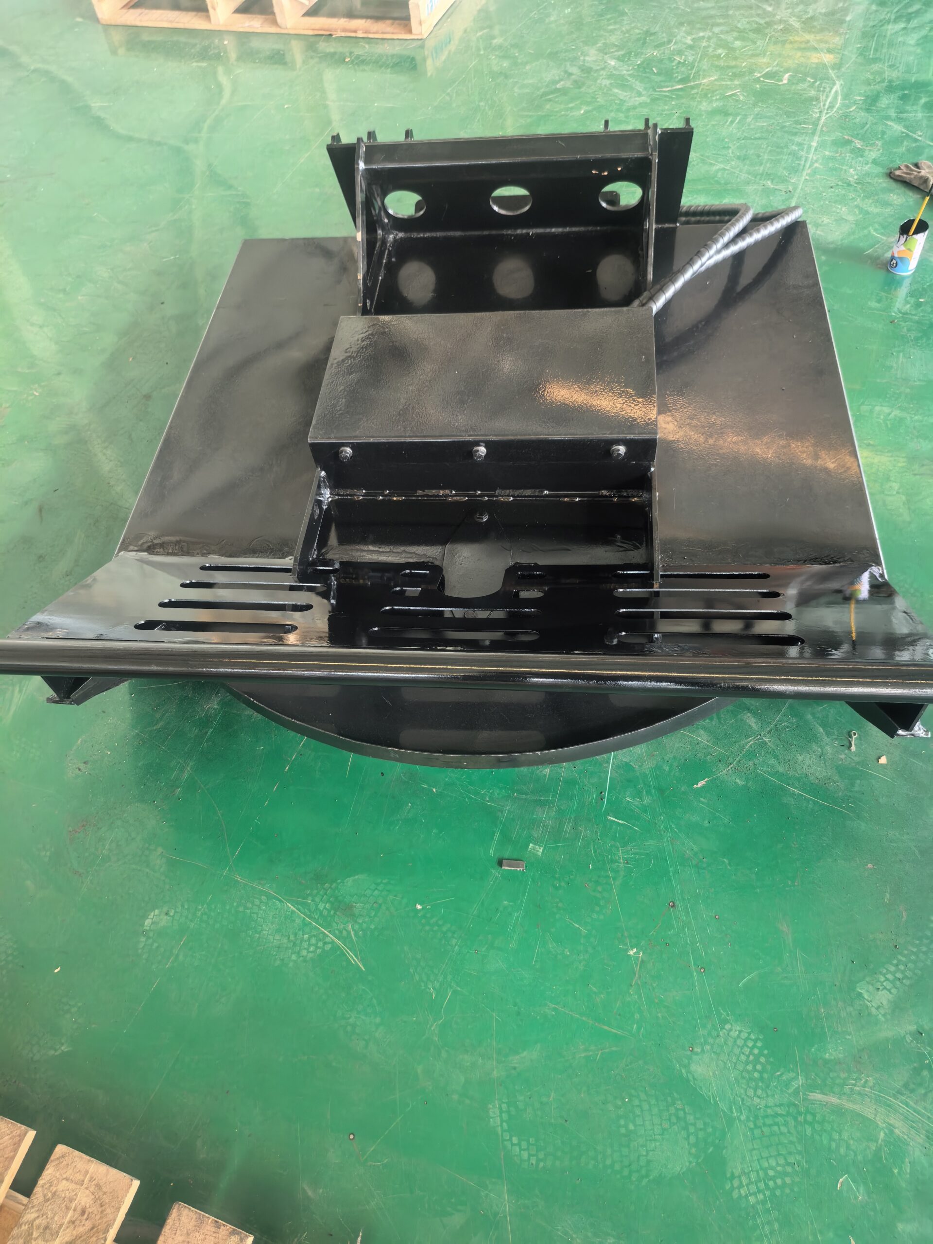 Black-painted metal industrial component with a sloped top plate and perforated base resting on a green workshop floor.