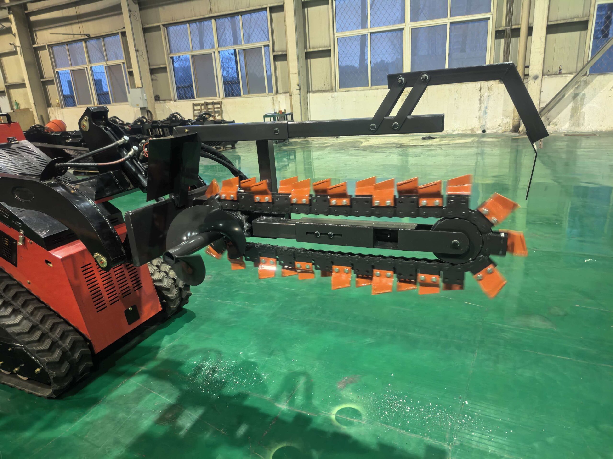 Industrial machine with a tracked base and long black arm featuring orange tines, in a factory setting with green floor and large windows.