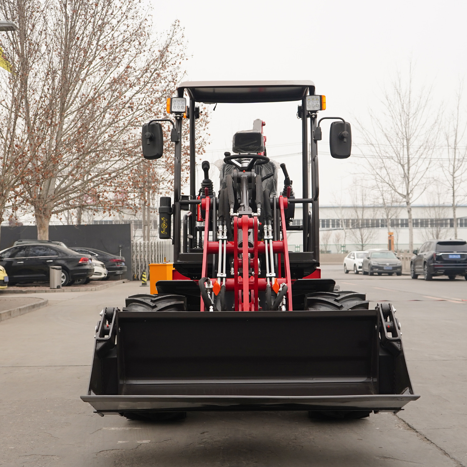 Back view of a heavy-duty tractor with red hydraulic arms and a large front loader bucket in a parking lot.