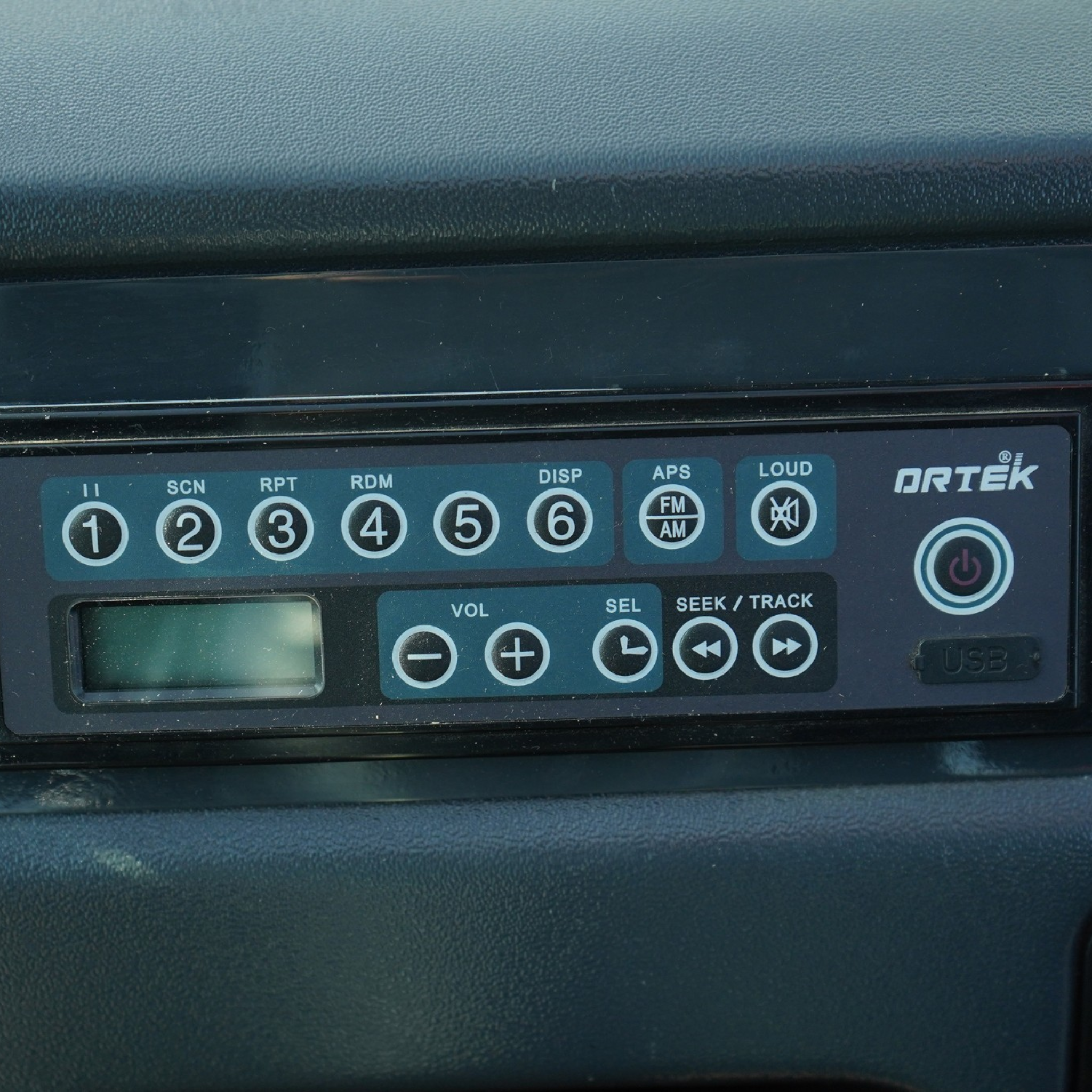Close-up of a car stereo control panel with numbered preset buttons 1–6 and auxiliary function labels like SCN, RPT, RDM, DISP, and APS on a blue surface.