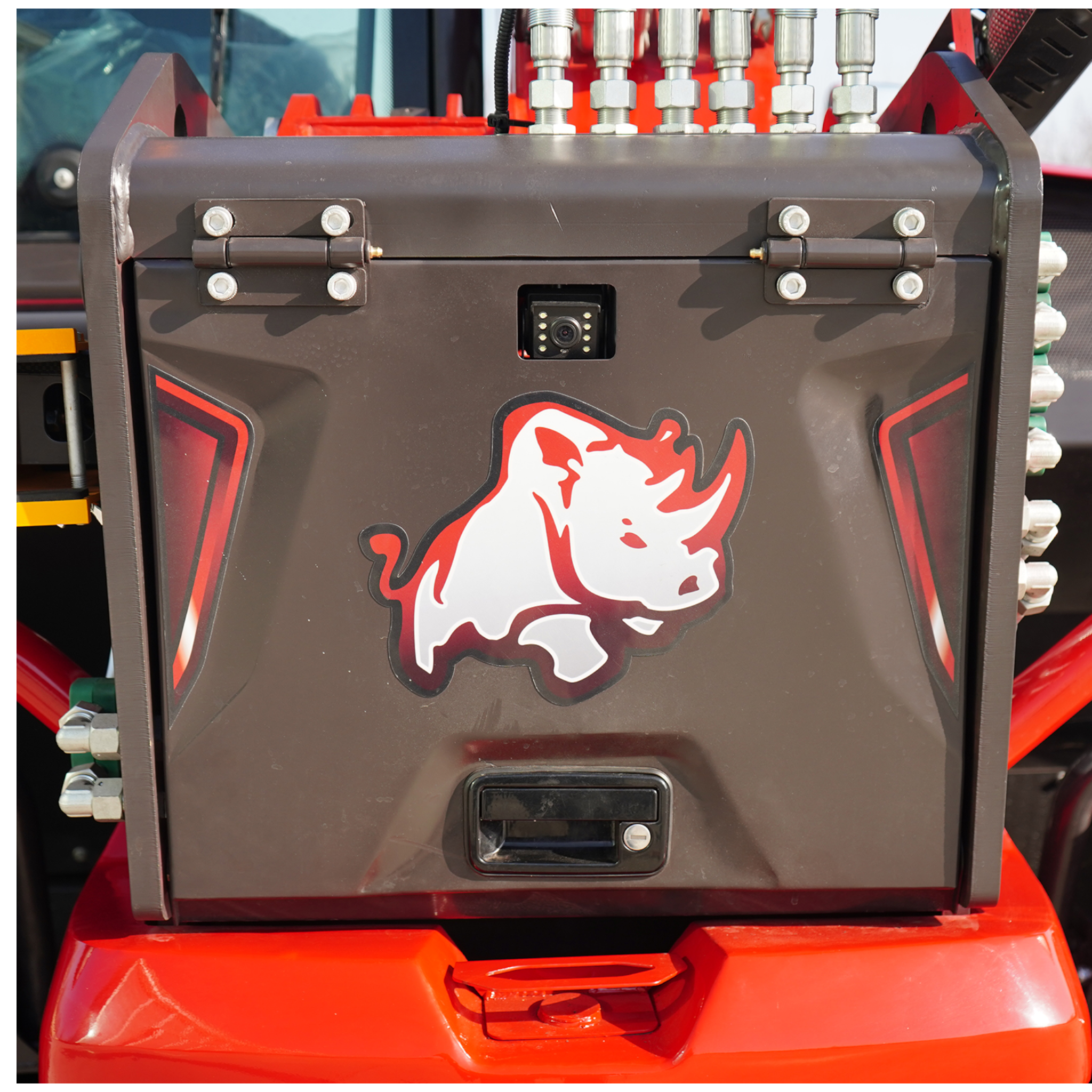 Close-up of a dark panel on a heavy vehicle with hydraulic fittings above and a small square camera in the center