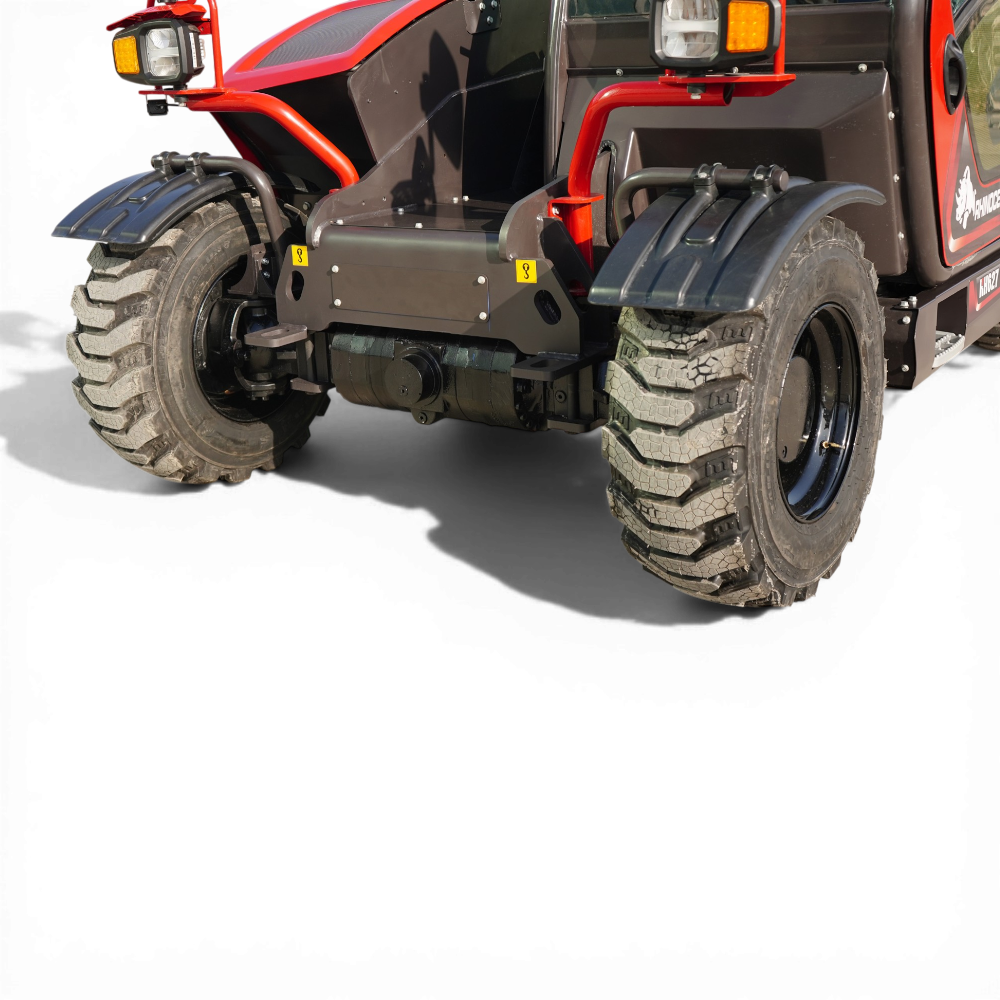 Close-up of a red heavy-duty vehicle's front wheel and chassis, showing a large off-road tire, red frame, yellow warning labels and orange headlights.