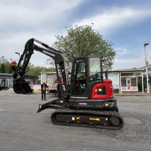 Red and black mini excavator with extended hydraulic arm in a construction yard, a worker in a yellow helmet nearby.