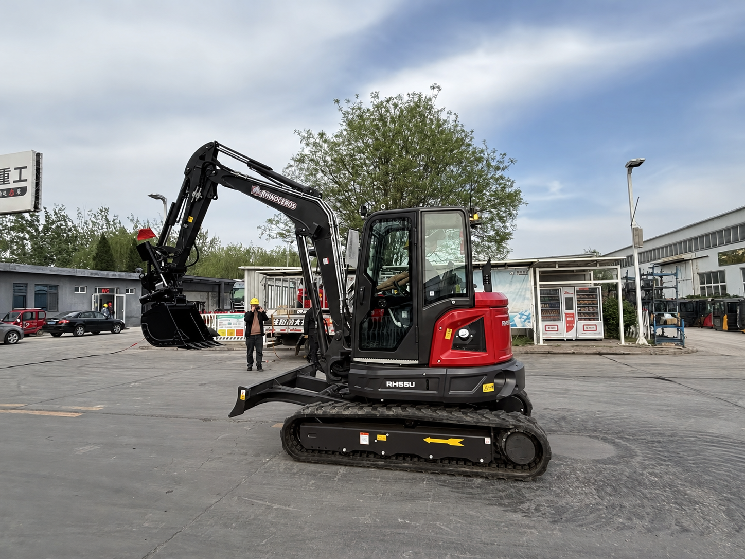 Red and black mini excavator with extended hydraulic arm in a construction yard, a worker in a yellow helmet nearby.