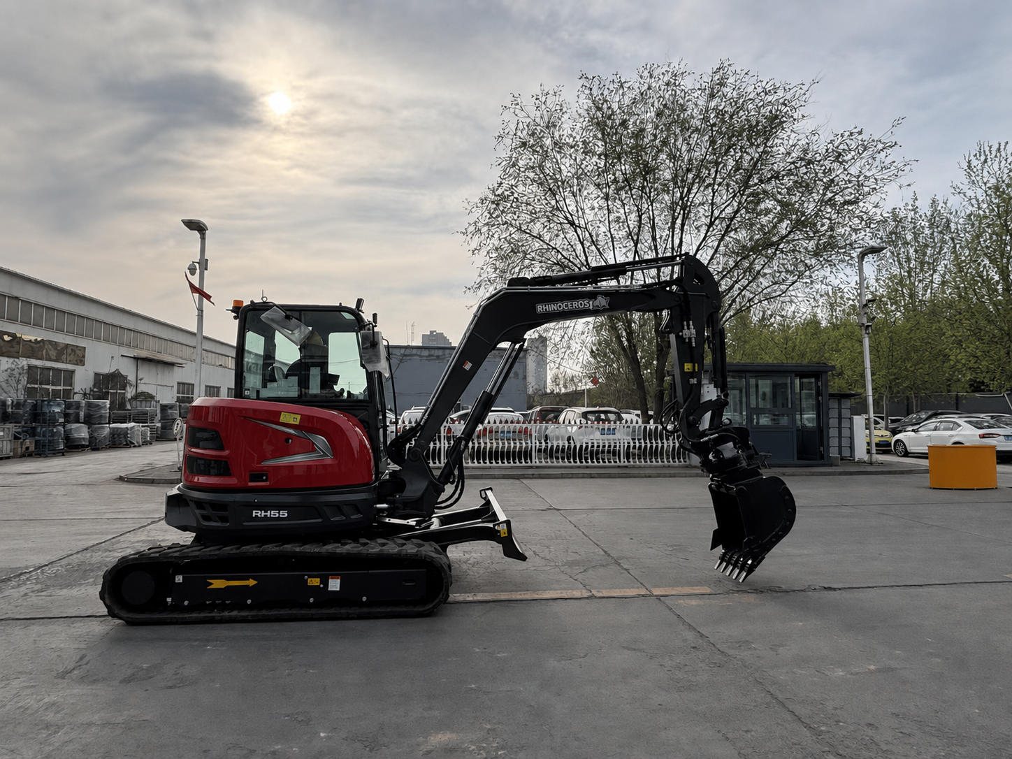 Red compact excavator with hydraulic arm extended and bucket toward the ground in an industrial yard, RH55 visible on the machine.